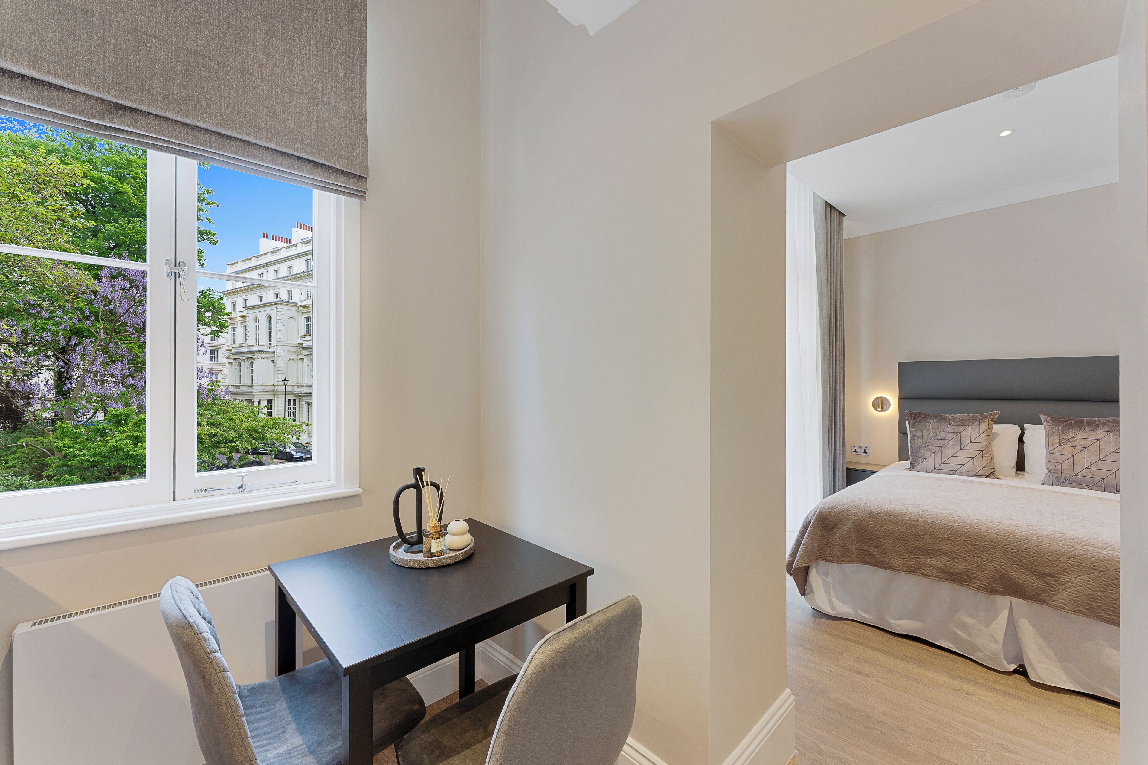 A hotel room with a bed, a table with a vase, and a view of a building and trees outside the window.
