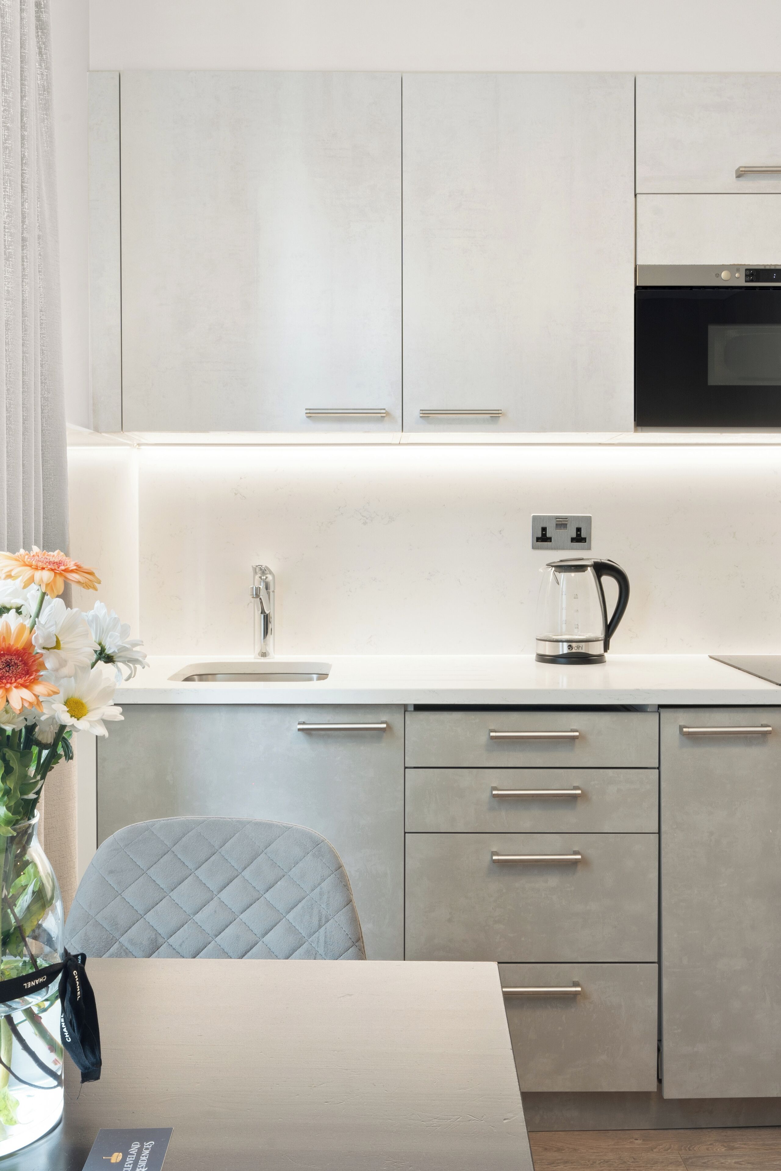 A modern kitchen with a built-in oven, a kettle, and a sink. There is a chair with a quilted backrest and a table with a vase of flowers.