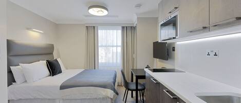 A modern hotel room with a bed, desk, and kitchenette.