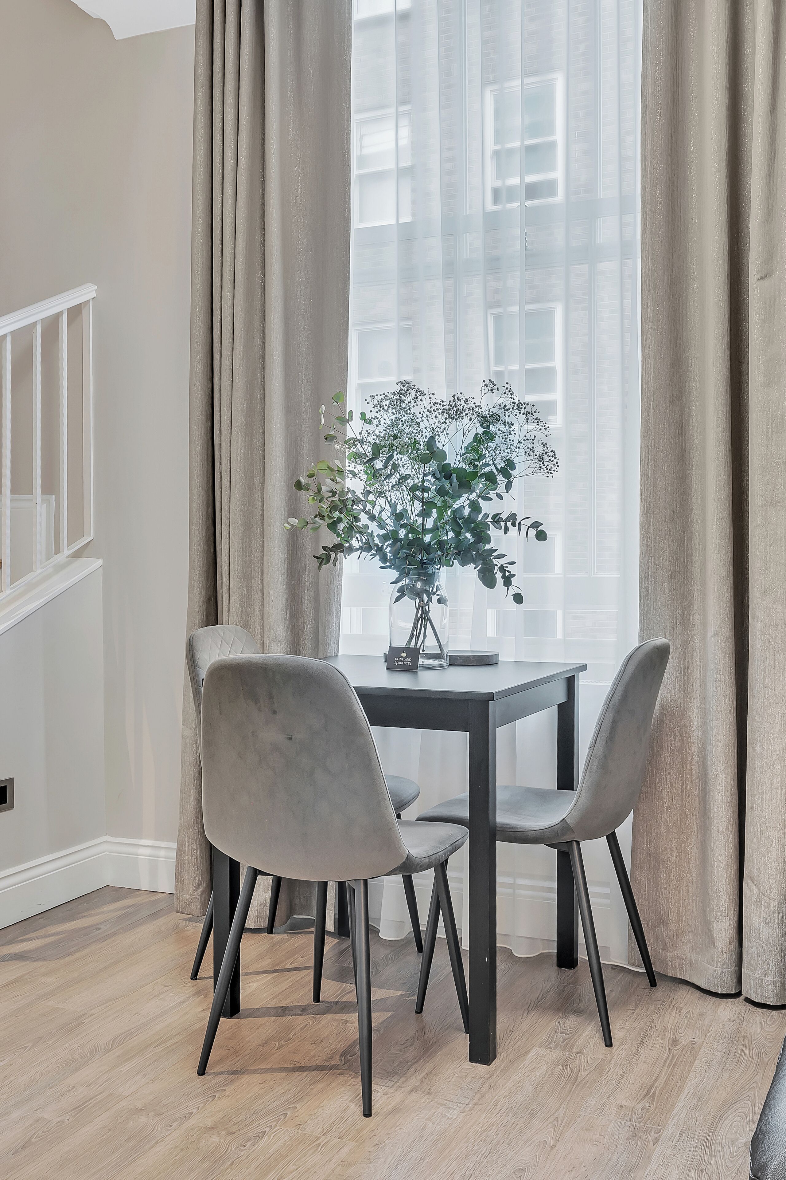 A dining area with a table and chairs, a vase with flowers, and a view through sheer curtains.