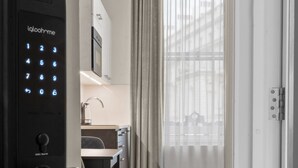 A modern hotel room with a built-in digital lock, a dining area with a table and chairs, and a large window with sheer curtains. - Cleveland Residences Paddington (London)