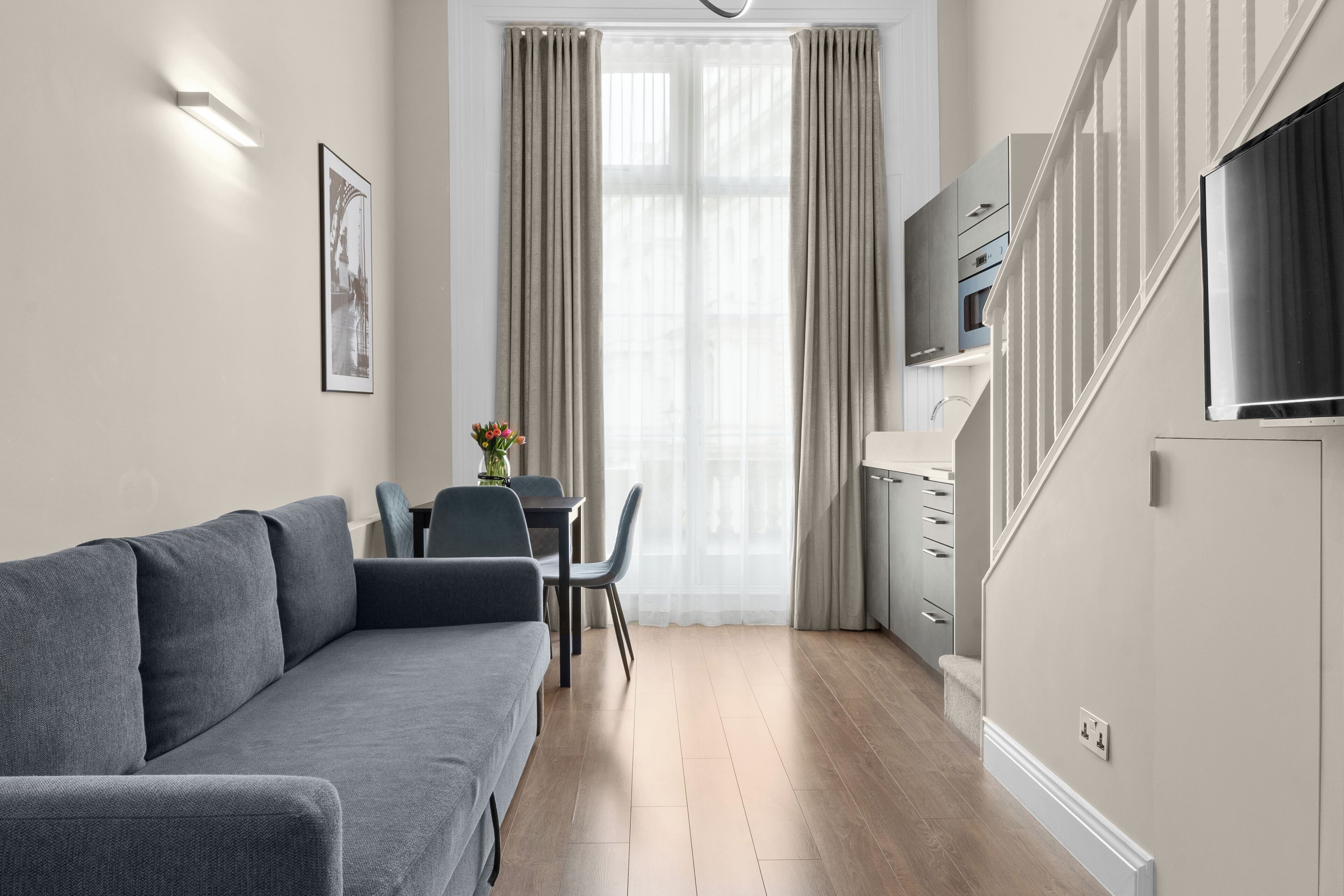 Un salon moderne avec un canapé gris, une table à manger avec des chaises, un téléviseur fixé au mur et un escalier menant à l’étage supérieur.