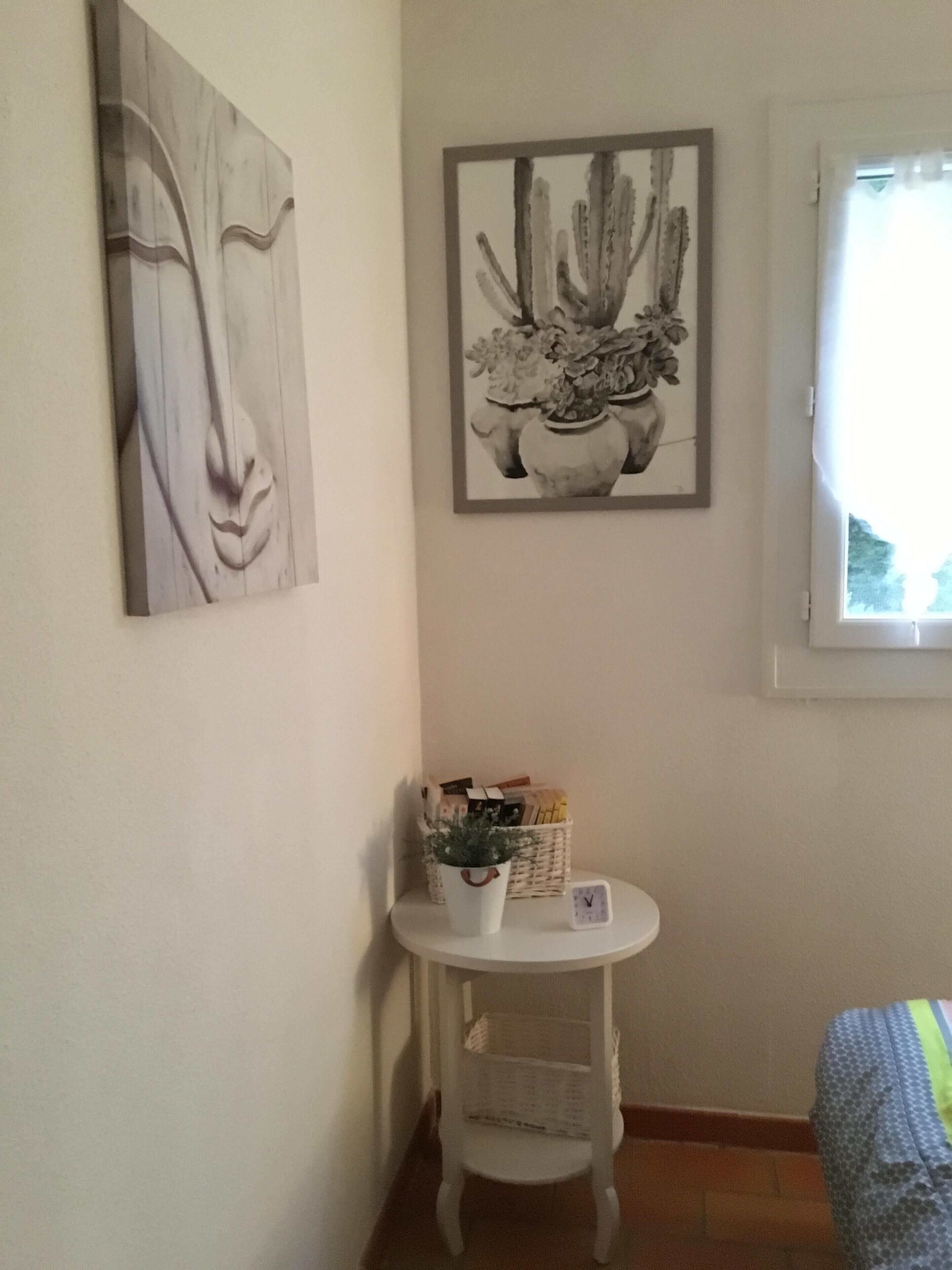 Una habitación pequeña con una cama, una mesa redonda blanca con un reloj, una cesta de libros y dos cuadros en la pared.