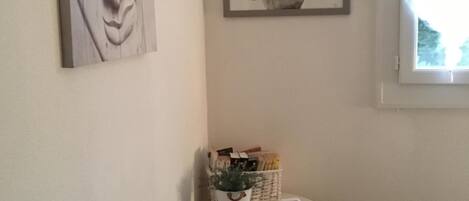 Una habitación pequeña con una cama, una mesa redonda blanca con un reloj, una cesta de libros y dos cuadros en la pared.