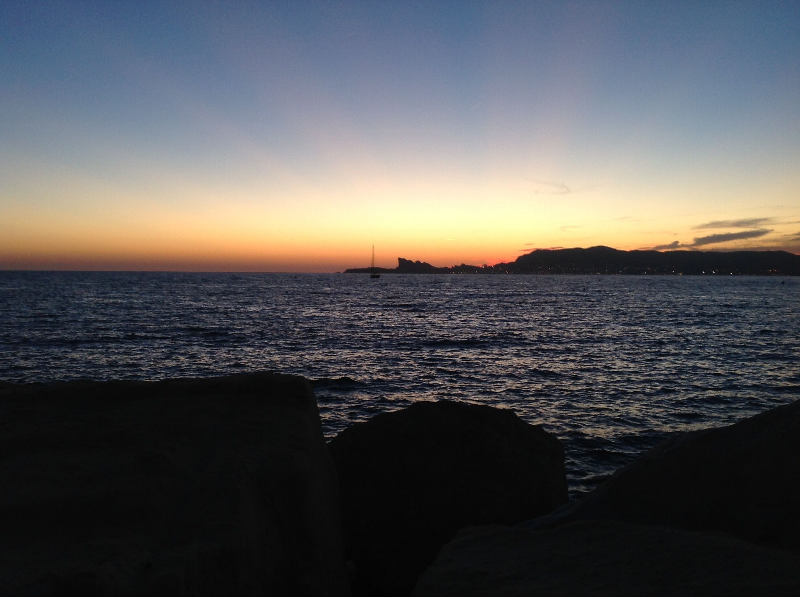 Ein Sonnenuntergang über einem Gewässer, mit einem Segelboot in der Ferne und einer felsigen Küste im Vordergrund.