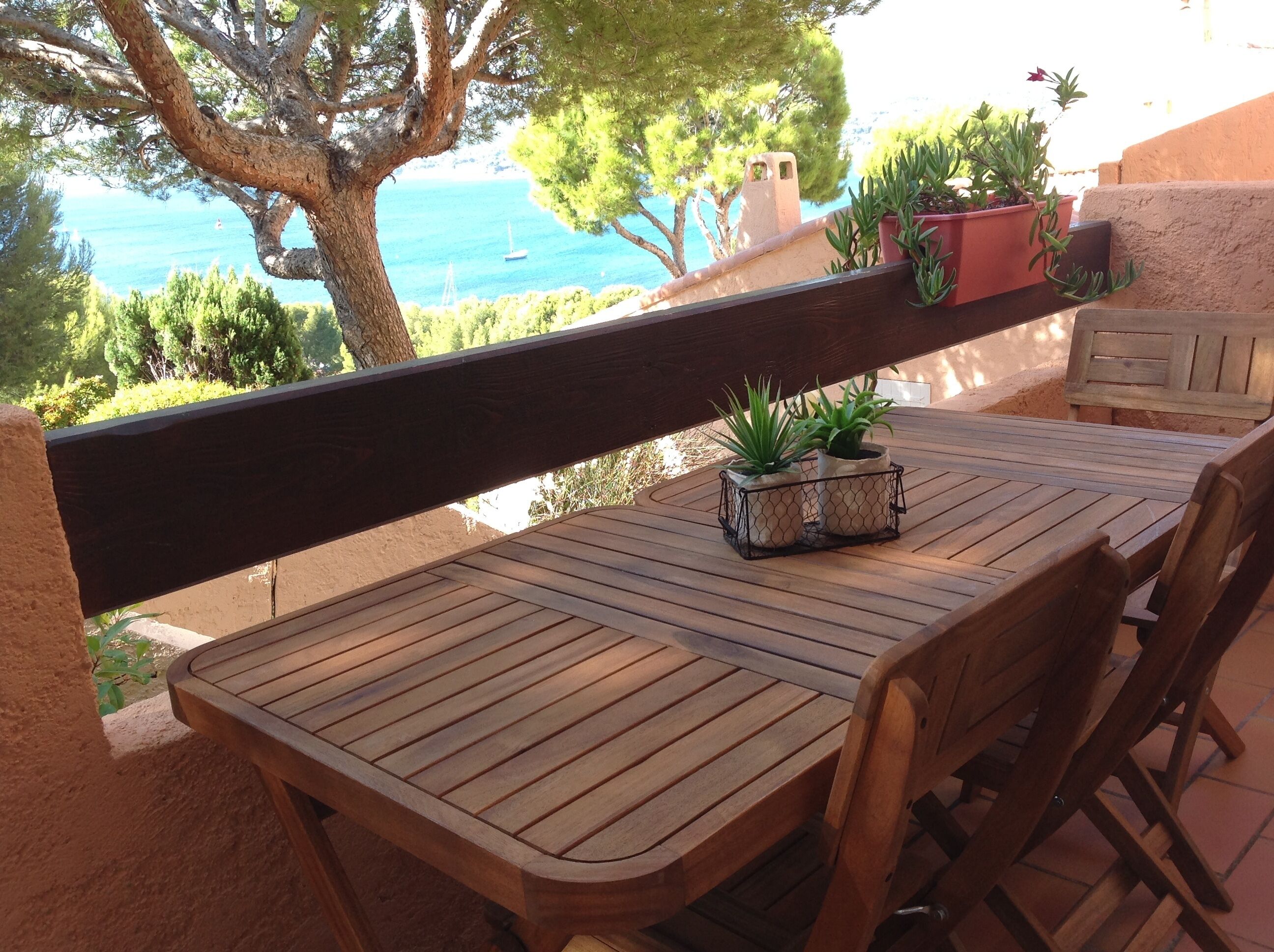 Een houten tafel met stoelen op een terras, met uitzicht op de kust, bomen en een heldere lucht.