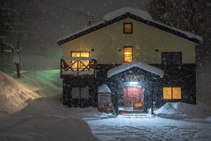 A snow-covered house with a lit entrance and a visible garage door. - Niseko Tabi-tsumugi Backpackers (Kutchan)