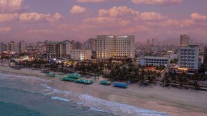 A coastal cityscape with a prominent hotel, beachfront with umbrellas, and palm trees. - DLG Hotel Danang (Da Nang)