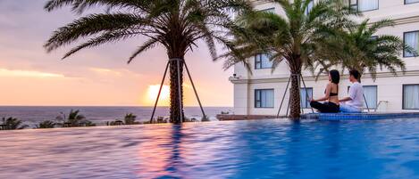 A poolside with two people sitting, palm trees, and a building with windows.