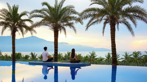 Two people meditating on an infinity pool with palm trees and a mountain view. - DLG Hotel Danang (Da Nang)
