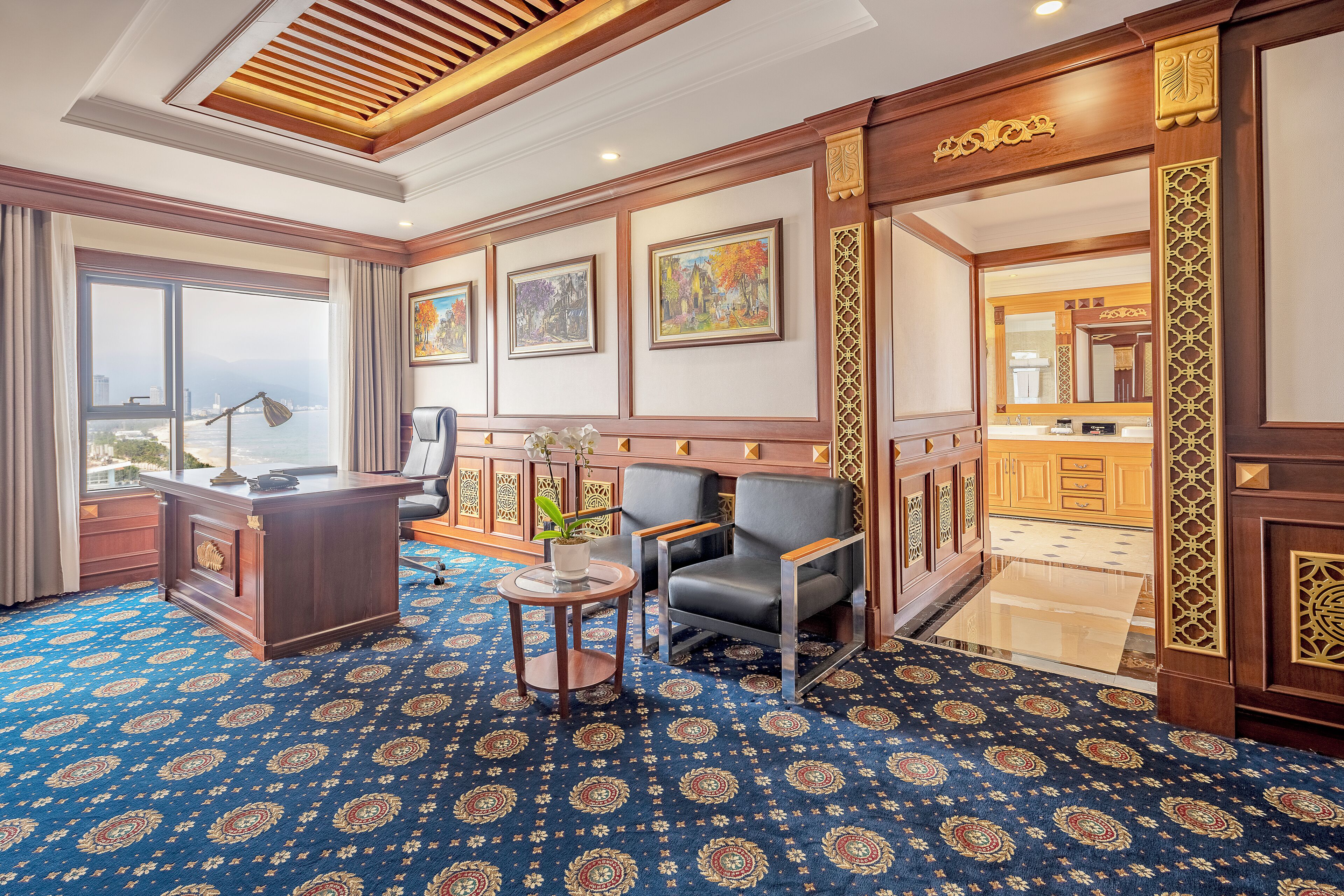 A hotel room with a large window, a desk, a chair, a small table with a plant, and a patterned carpet.