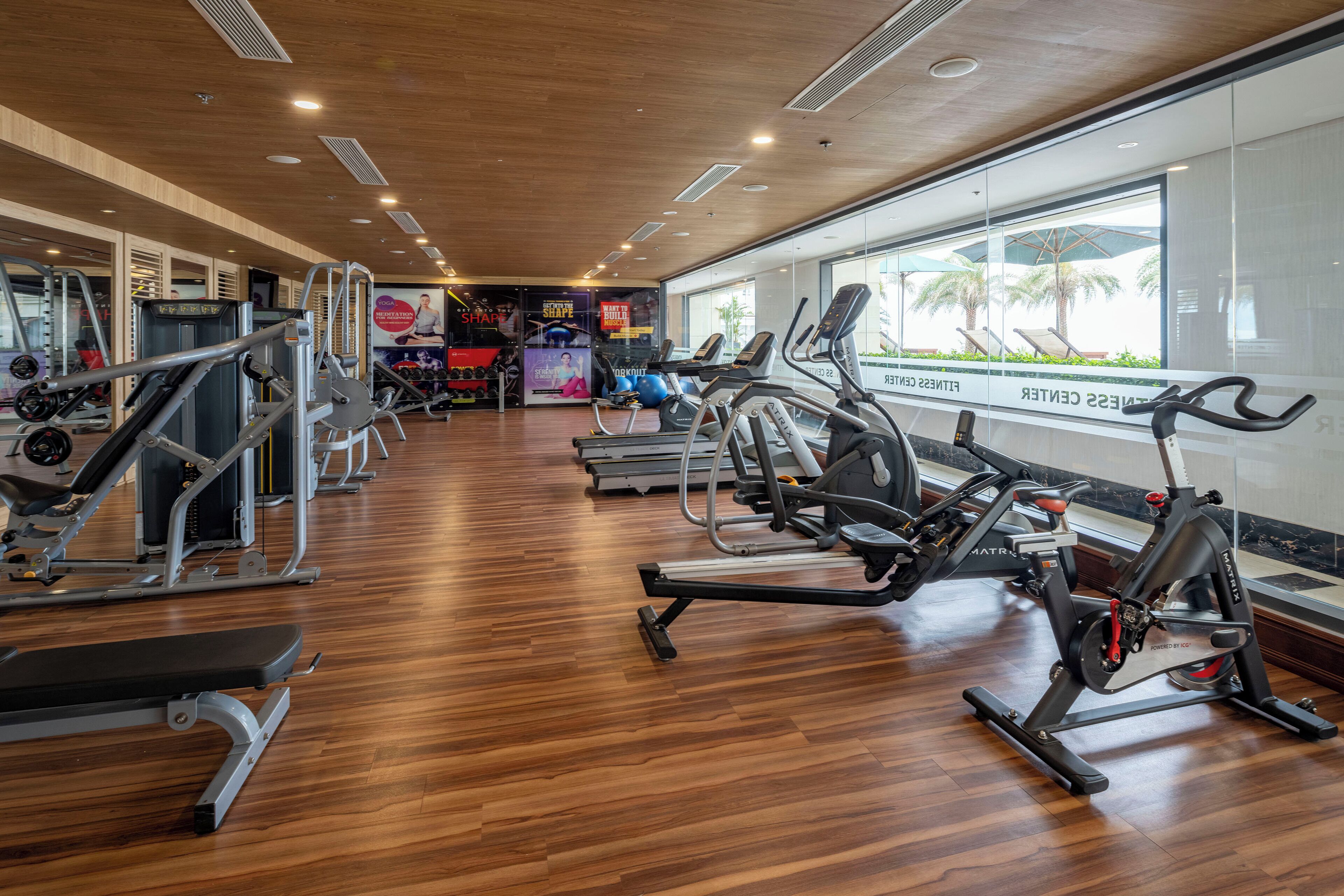 Een fitnessruimte met diverse trainingsapparaten, grote ramen en een houten vloer.