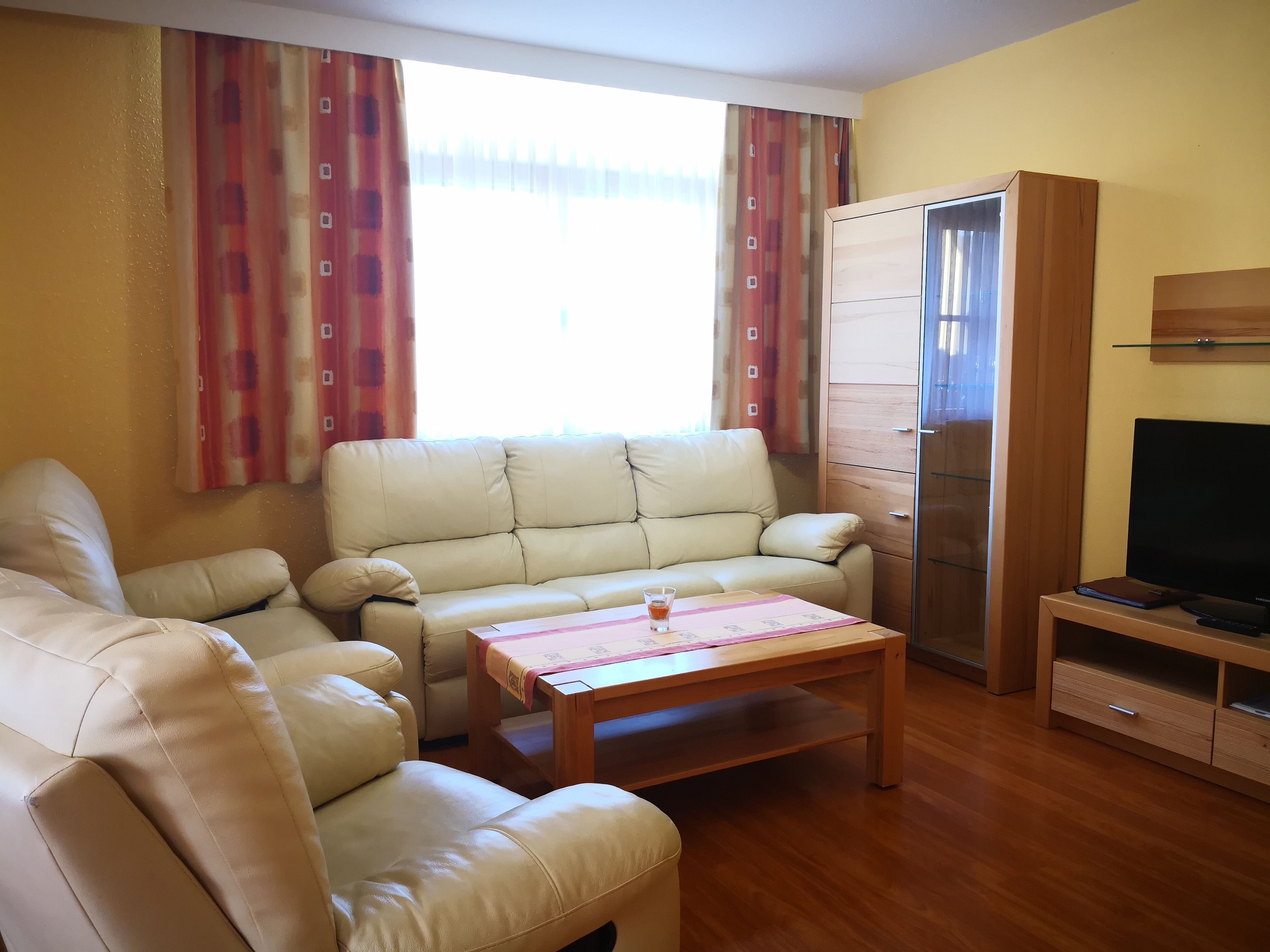 Uma sala de estar com um sofá bege, uma mesa de centro de madeira, uma TV em um suporte de madeira e um guarda-roupa.
