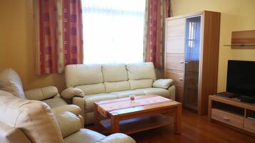 Uma sala de estar com um sofá bege, uma mesa de centro de madeira, uma TV em um suporte de madeira e um guarda-roupa.