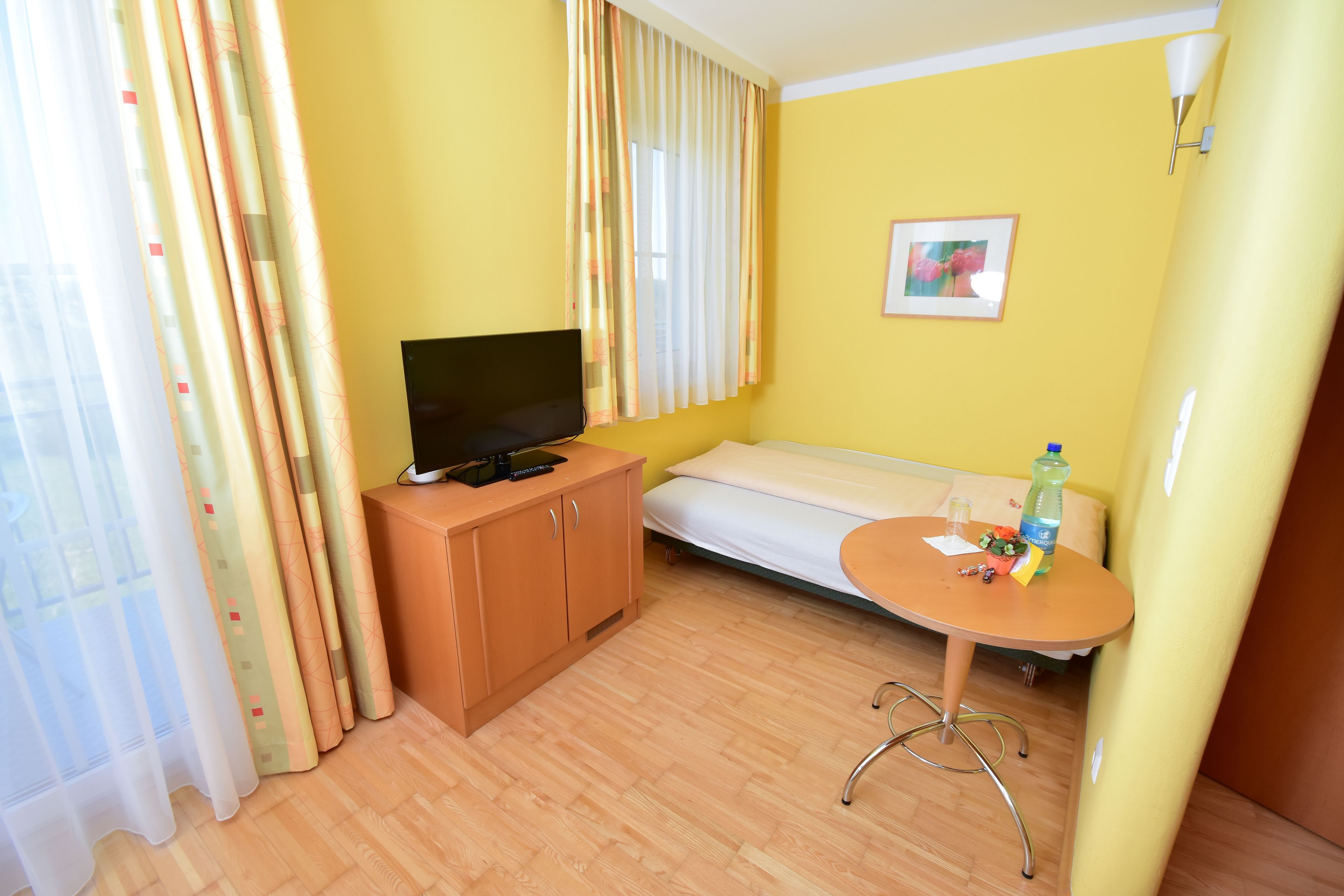 Una habitación con una cama, un televisor sobre un mueble de madera, una mesita redonda con una botella y un cuadro en la pared.