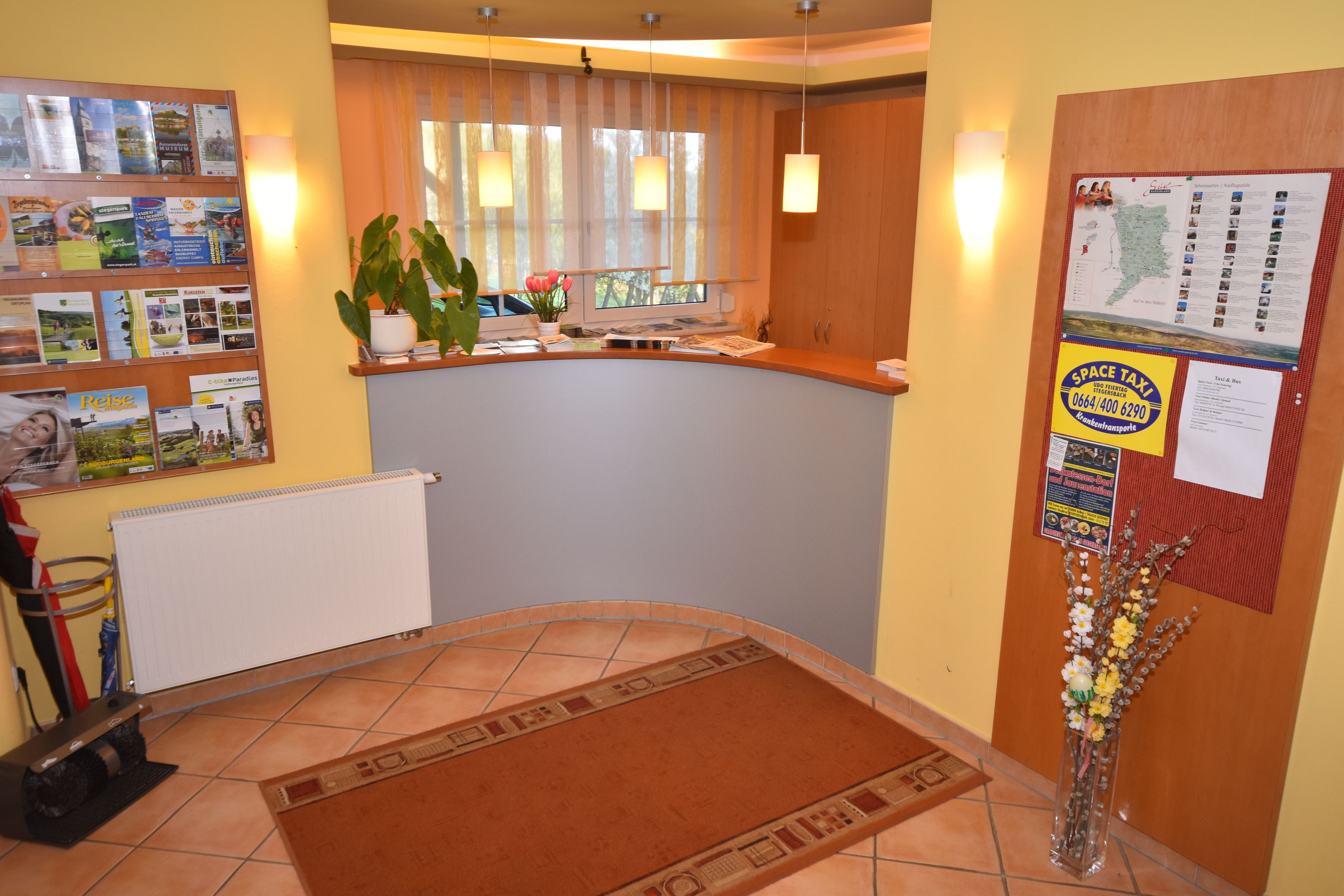 A reception area with a counter, a potted plant, a bulletin board with maps and notices, and a tiled floor.