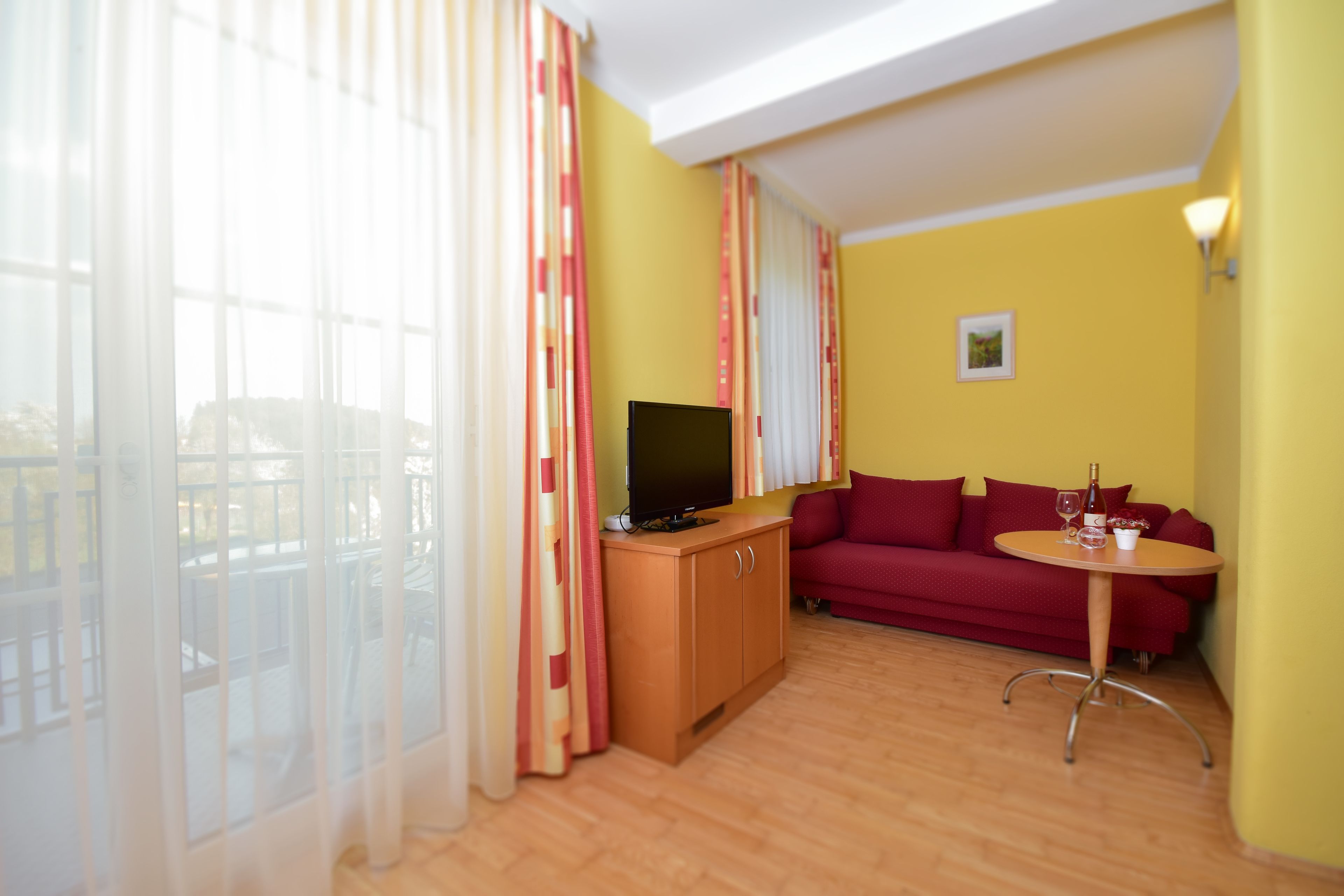 A living room with a sofa, a small table with a bottle and glass, a TV on a wooden cabinet, and large windows with sheer curtains.