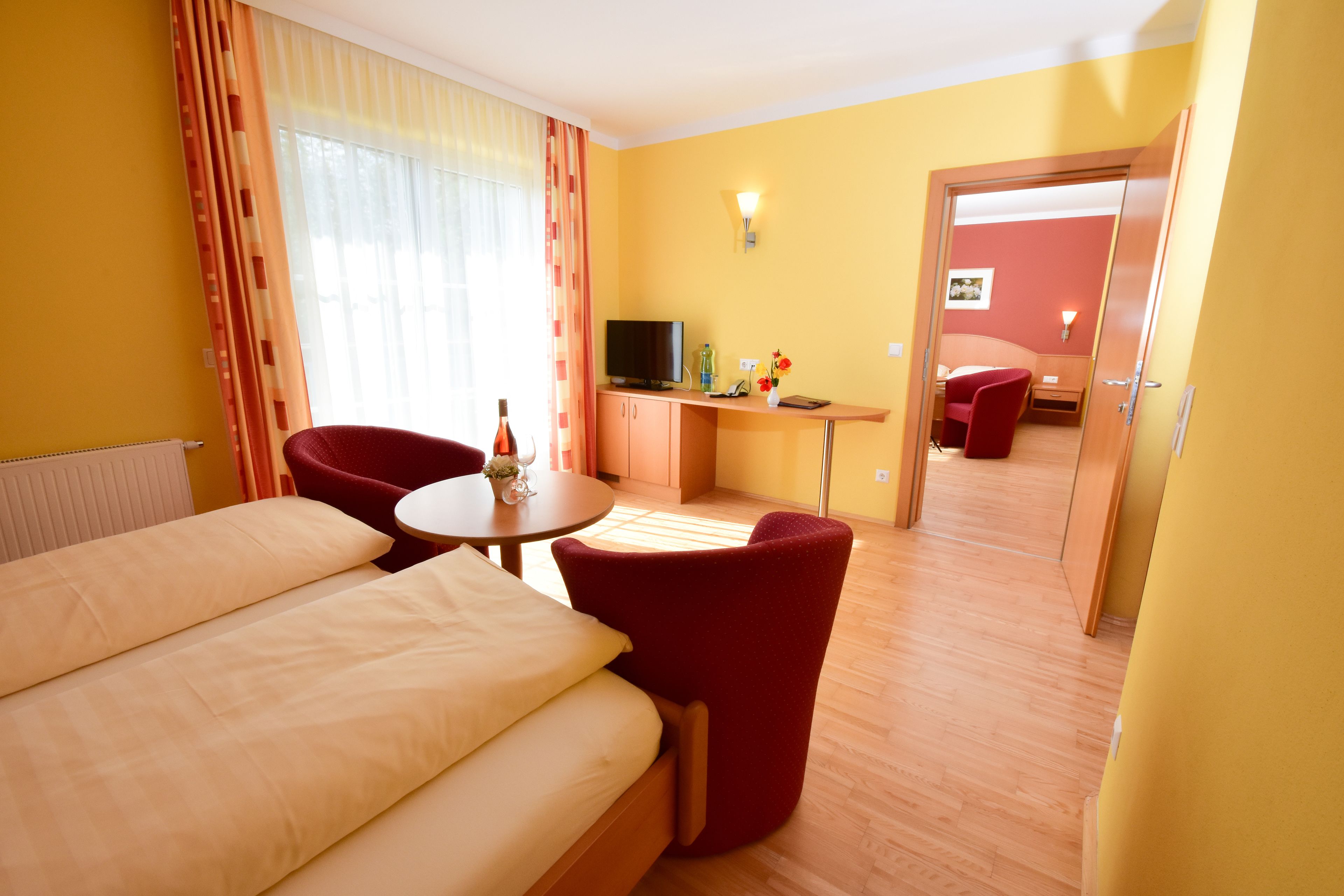 A hotel room with a bed, a desk with a TV, a chair, and a small table with a vase.