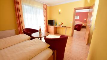 A hotel room with a bed, a desk with a TV, a chair, and a small table with a vase.