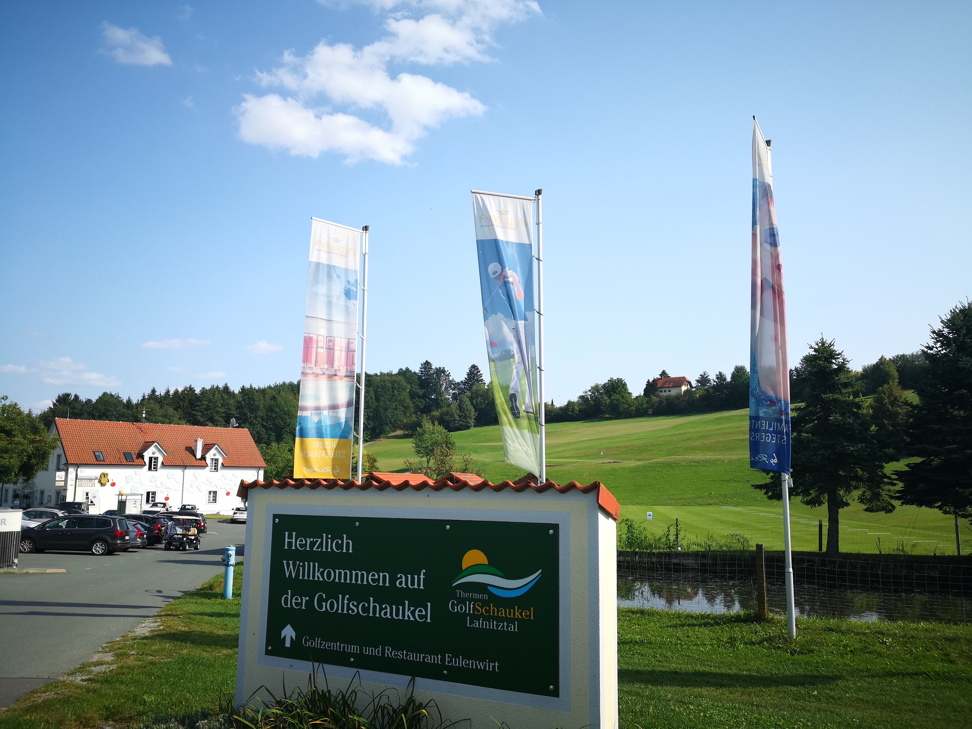 Un letrero da la bienvenida a visitantes a un club de golf, con un campo verde y un edificio al fondo.