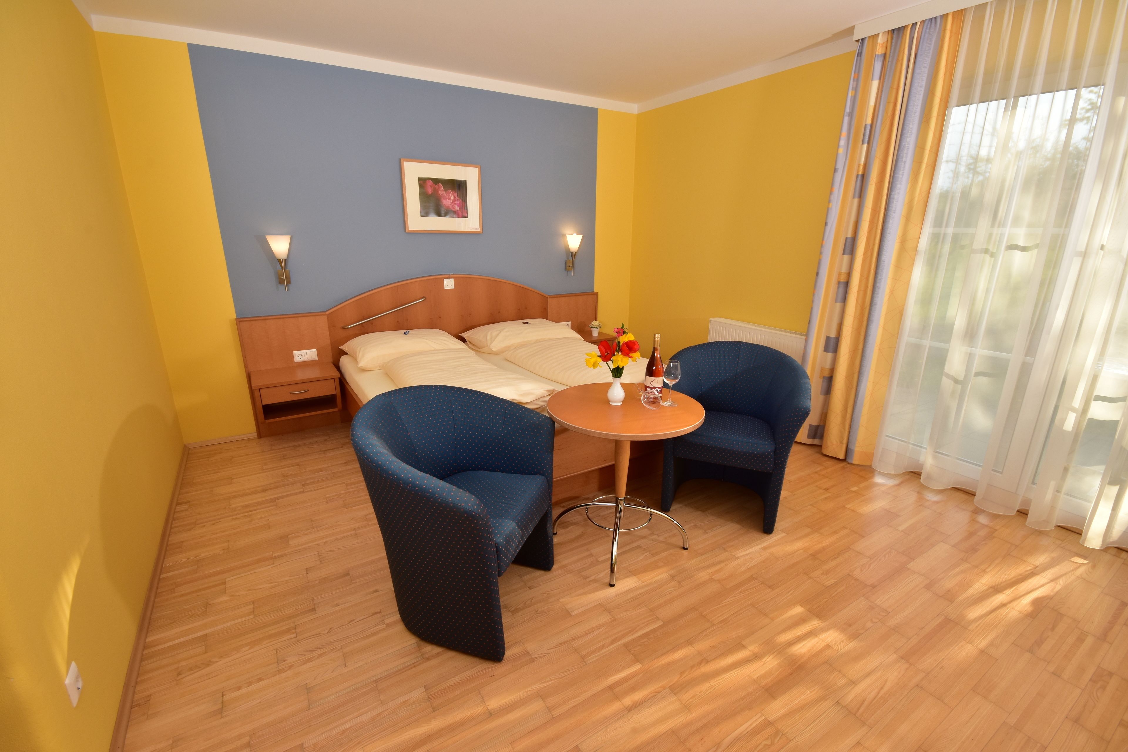 A hotel room with a bed, two armchairs, a small table with a vase of flowers, and a picture on the wall.