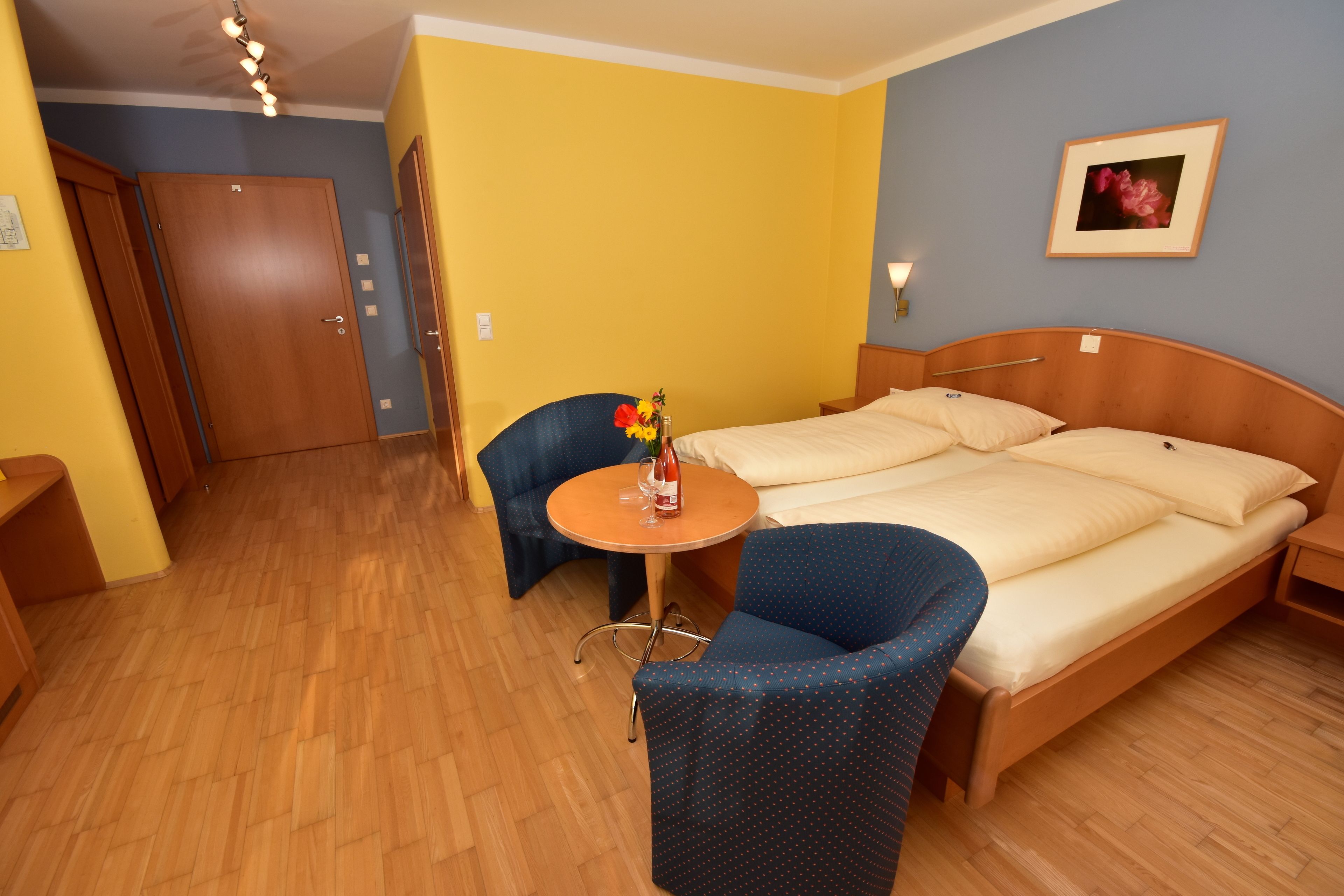 Une chambre d’hôtel avec un lit en bois, une petite table ronde ornée d’un vase de fleurs, une chaise bleue et une armoire en bois.