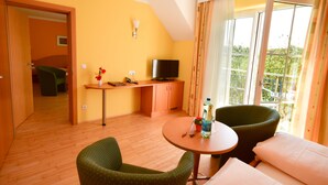 A room with a wooden floor, a small round table, two green chairs, a sofa, a TV, a small cabinet, and a window with curtains. - Golf & Therme Sonnenpension Hotel Garni (Burgauberg-Neudauberg)
