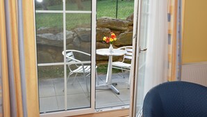 A room with a sliding glass door leading to an outdoor patio area with a white metal table and chairs, a vase with flowers, and a stone wall. - Golf & Therme Sonnenpension Hotel Garni (Burgauberg-Neudauberg)