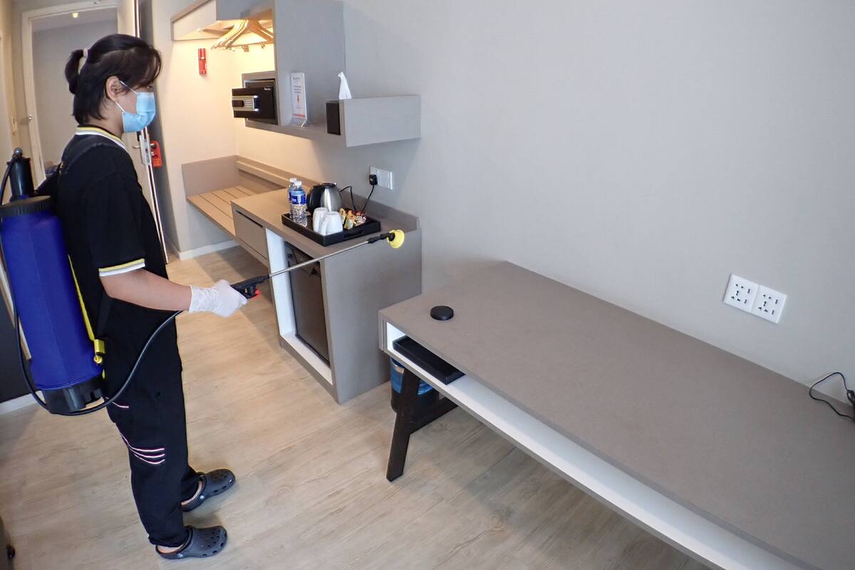 Una persona usando un dispositivo de luz ultravioleta en una habitación con una mesa larga, una pequeña área de cocina y una puerta que conduce a otra habitación.