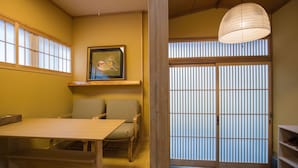 Una habitación japonesa tradicional con suelo de tatami, una mesa de madera baja y sillas. Cuenta con una puerta corredera de shoji, un farolillo de papel colgante y un cuadro en la pared.
