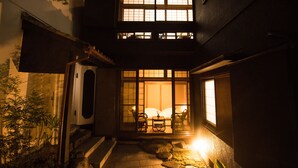 Un edificio tradicional japonés con fachada de madera, un sendero de piedra que lleva a un área de descanso con una mesa y sillas, y vistas del interior a través de amplias puertas correderas.