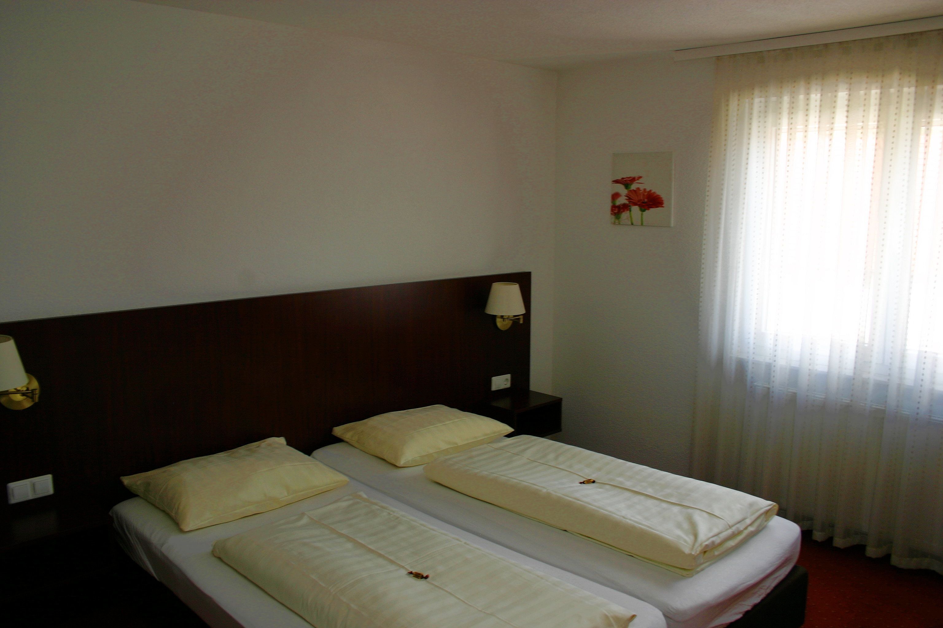 Een hotelkamer met twee bedden, een houten hoofdeinde, een nachtkastje met een lamp en een raam met dunne gordijnen.