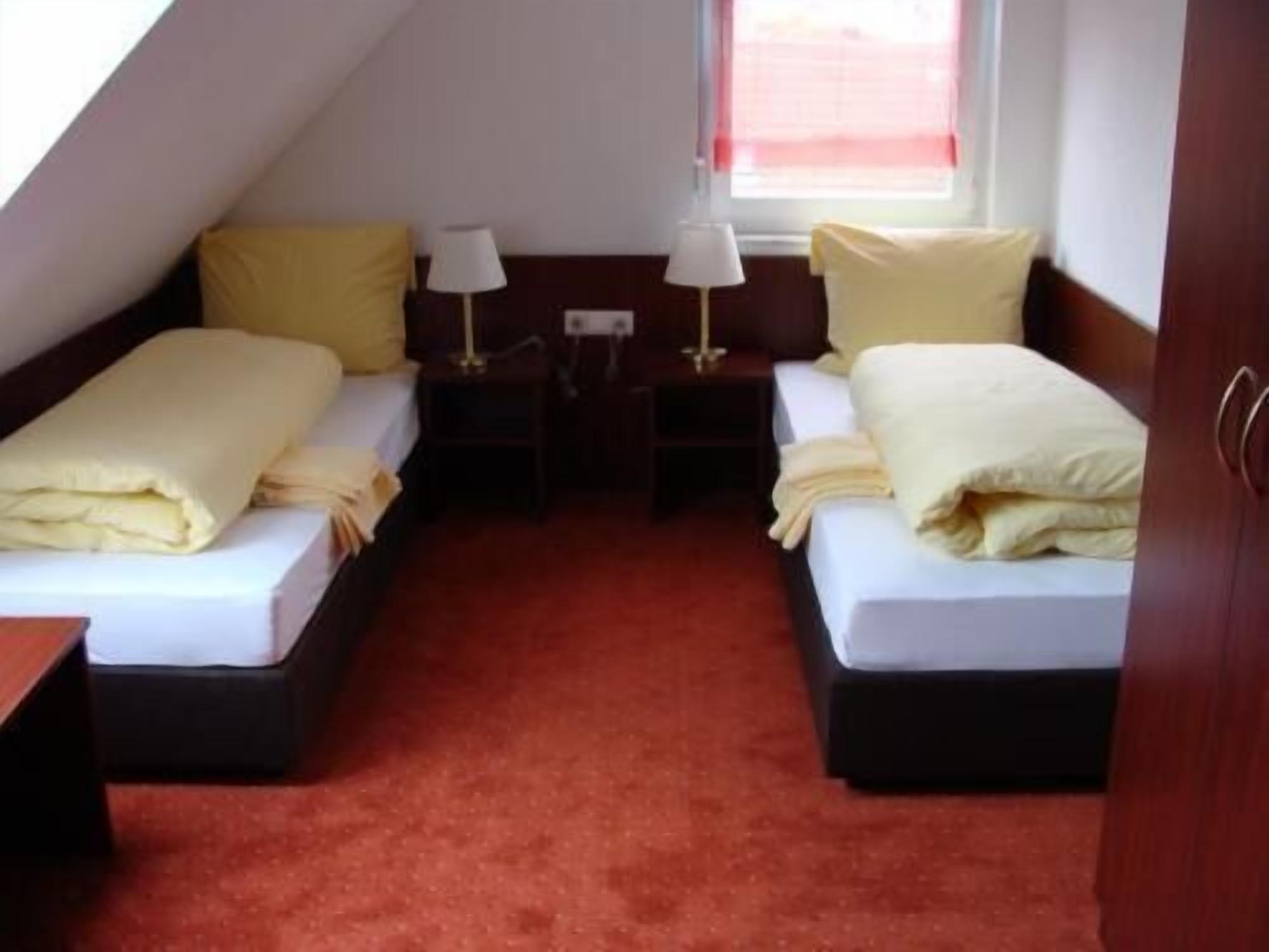 Een kleine, knusse kamer met twee eenpersoonsbedden, elk met een kussen en een lamp. De kamer heeft een rood tapijt en een raam met een rode gordijn.