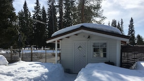 A small, white cabin with a hipped roof and a door with a diamond pattern. It is surrounded by snow and has a window with a view of the surrounding forest. - ♥ Seaside cottage ♥ Boat fishing snowmobile running (Luleå Ö)