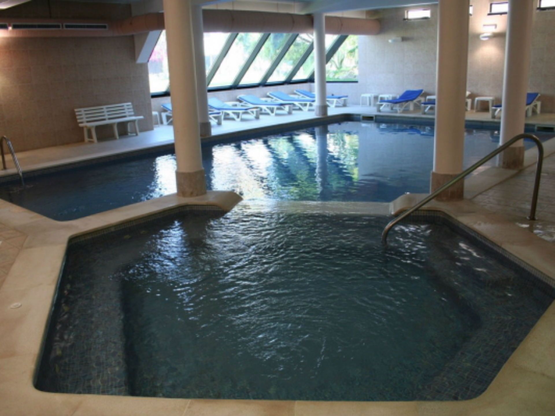 Une piscine intérieure à l’eau claire, entourée de chaises longues et d’un banc.