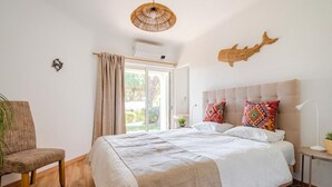 Une chambre à coucher comprenant un lit, une chaise, une table de chevet, une lampe, un élément de décoration murale et une fenêtre avec des rideaux.