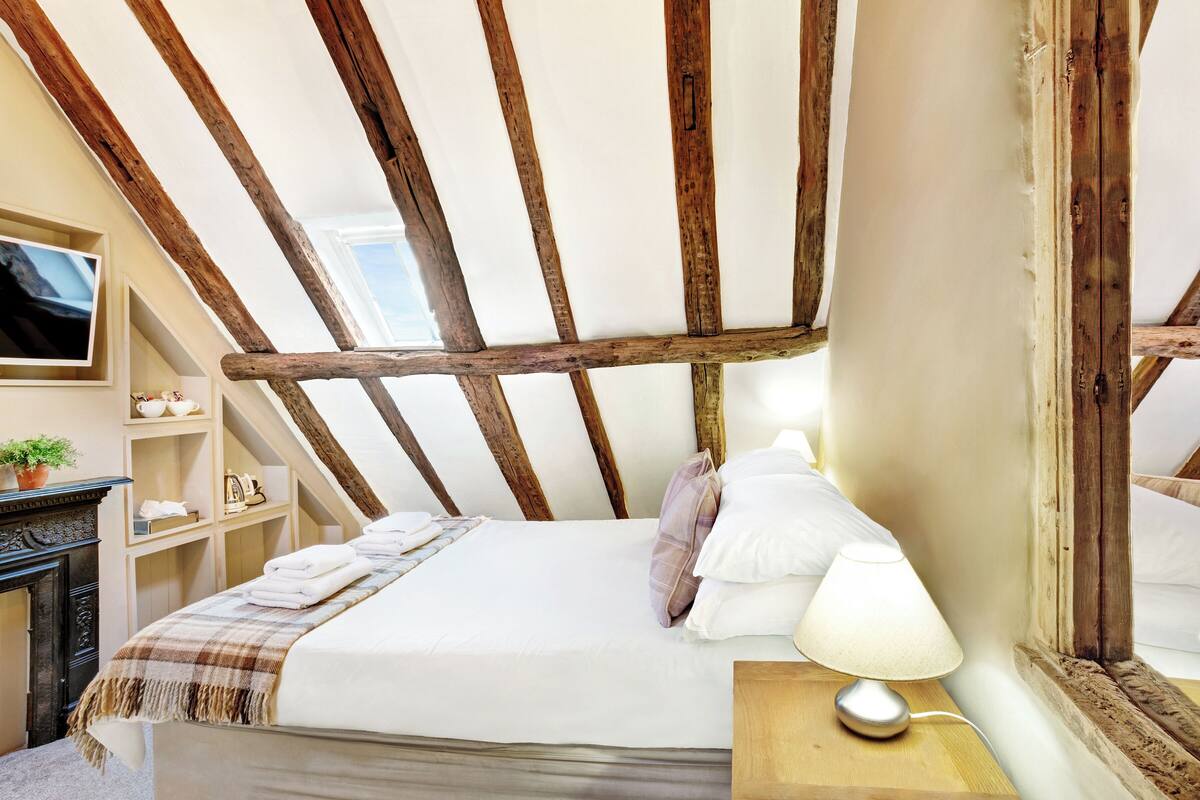 A bedroom with a sloped ceiling, wooden beams, a bed with white linens, a bedside table with a lamp, and a TV mounted on the wall.