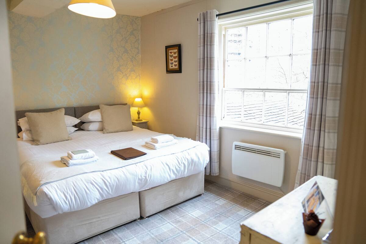 A neatly made bed with pillows and a bedside table with a lamp and a book. A window with curtains is visible, and a radiator is mounted on the wall.