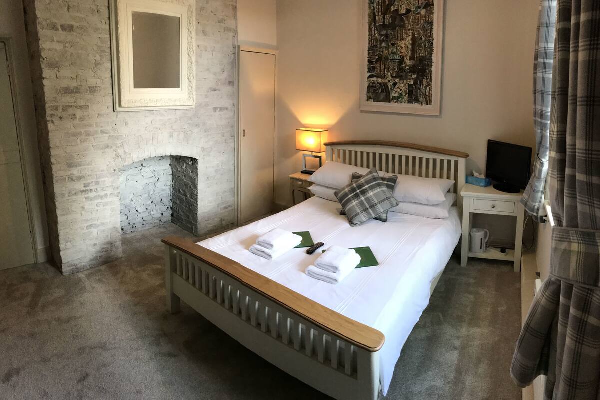 A neatly made bed with white linens and a plaid pillow, a bedside lamp, a television on a nightstand, and a painting on the wall.