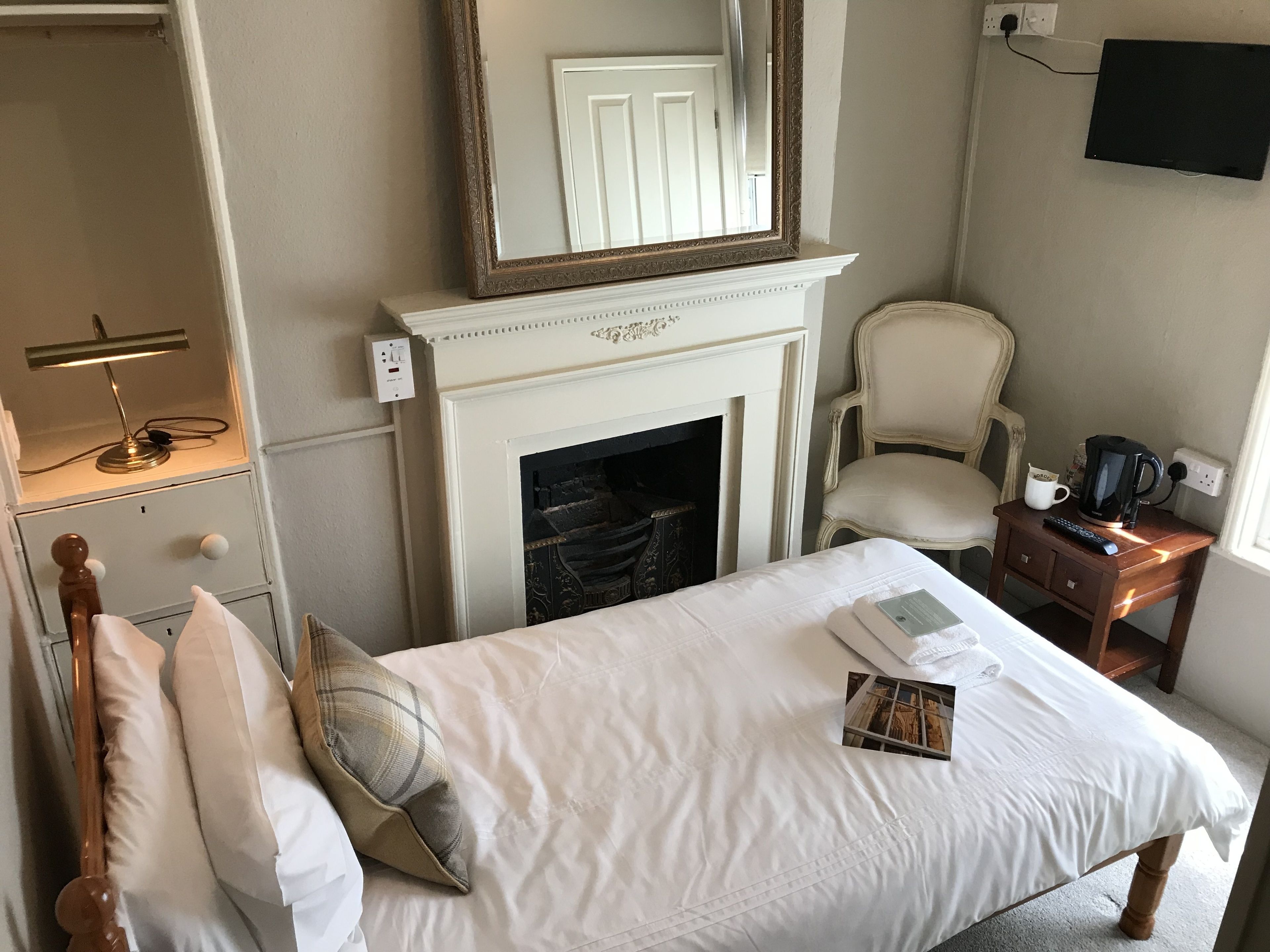 Une chambre d’hôtel équipée d’un lit, d’une chaise, d’une petite table avec une bouilloire, d’une cheminée et d’un téléviseur fixé au mur.