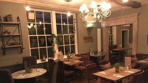 A cozy restaurant interior with wooden tables and chairs, a chandelier, and a large window with decorative elements. - Galtres Lodge Hotel (York)