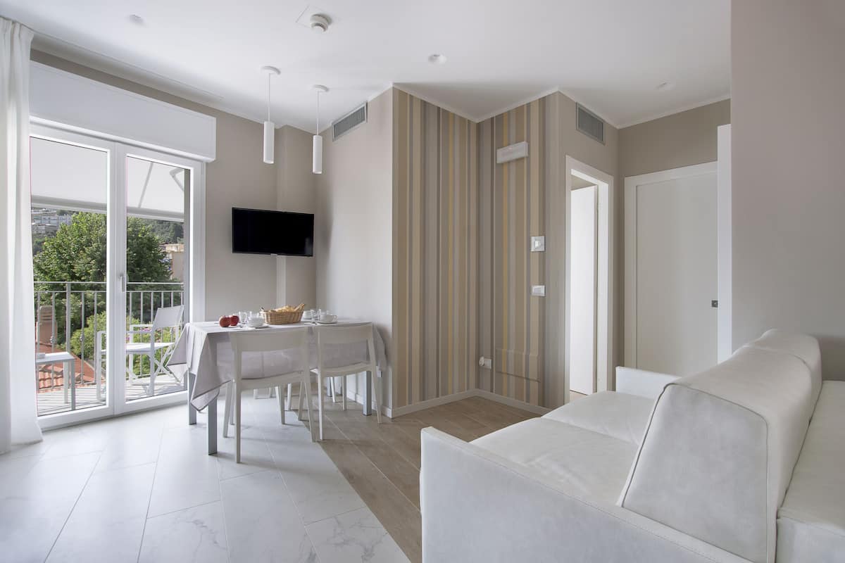 A modern living room with a white sofa, a dining area with a table and chairs, a TV mounted on the wall, and a sliding glass door leading to an outdoor area.