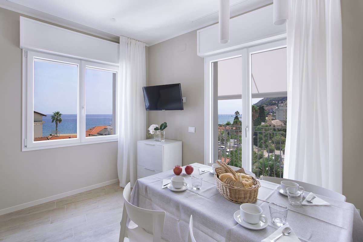 A dining area with a table set for breakfast, a TV mounted on the wall, and a balcony with a view of the sea.