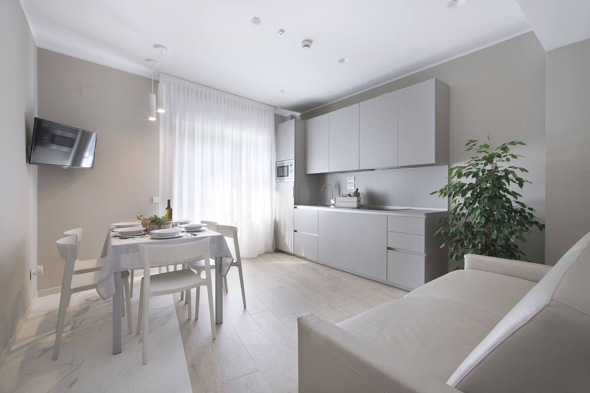 A modern kitchen with white cabinets, a dining area with a table and chairs, and a flat-screen TV mounted on the wall.