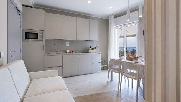 Une cuisine moderne dotée d’un four à micro-ondes encastré, un coin repas avec une table et des chaises blanches, et une vue sur la mer grâce à de grandes fenêtres.
