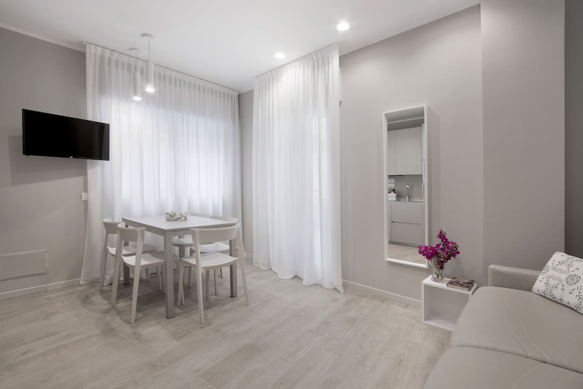 A modern, minimalist room with a dining table, chairs, a sofa, a TV, and a small side table with a vase of flowers.