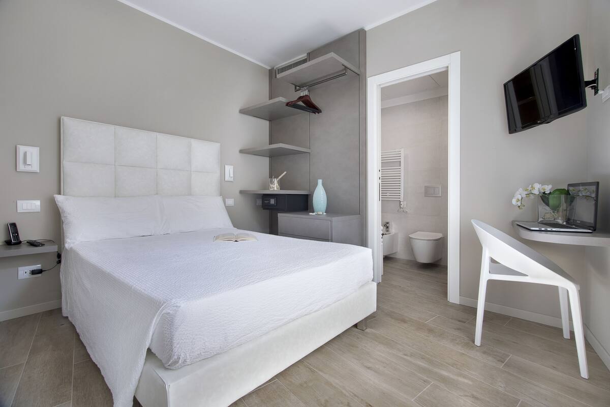A modern bedroom with a large bed, a bedside table, a TV mounted on the wall, and a small desk with a chair.