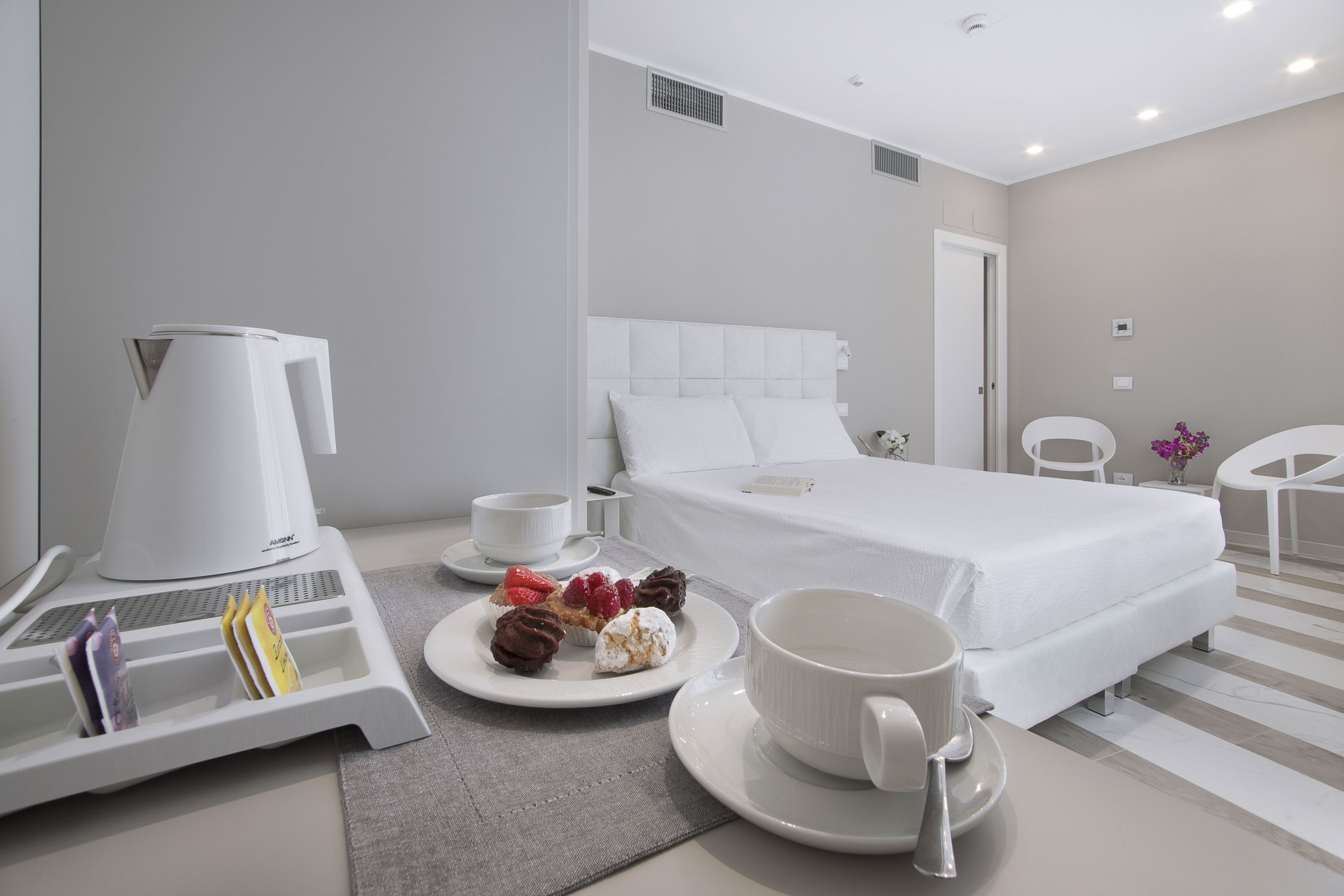 Quarto de hotel com uma cama, uma máquina de café, uma bandeja com pastéis e uma chávena sobre uma mesa.
