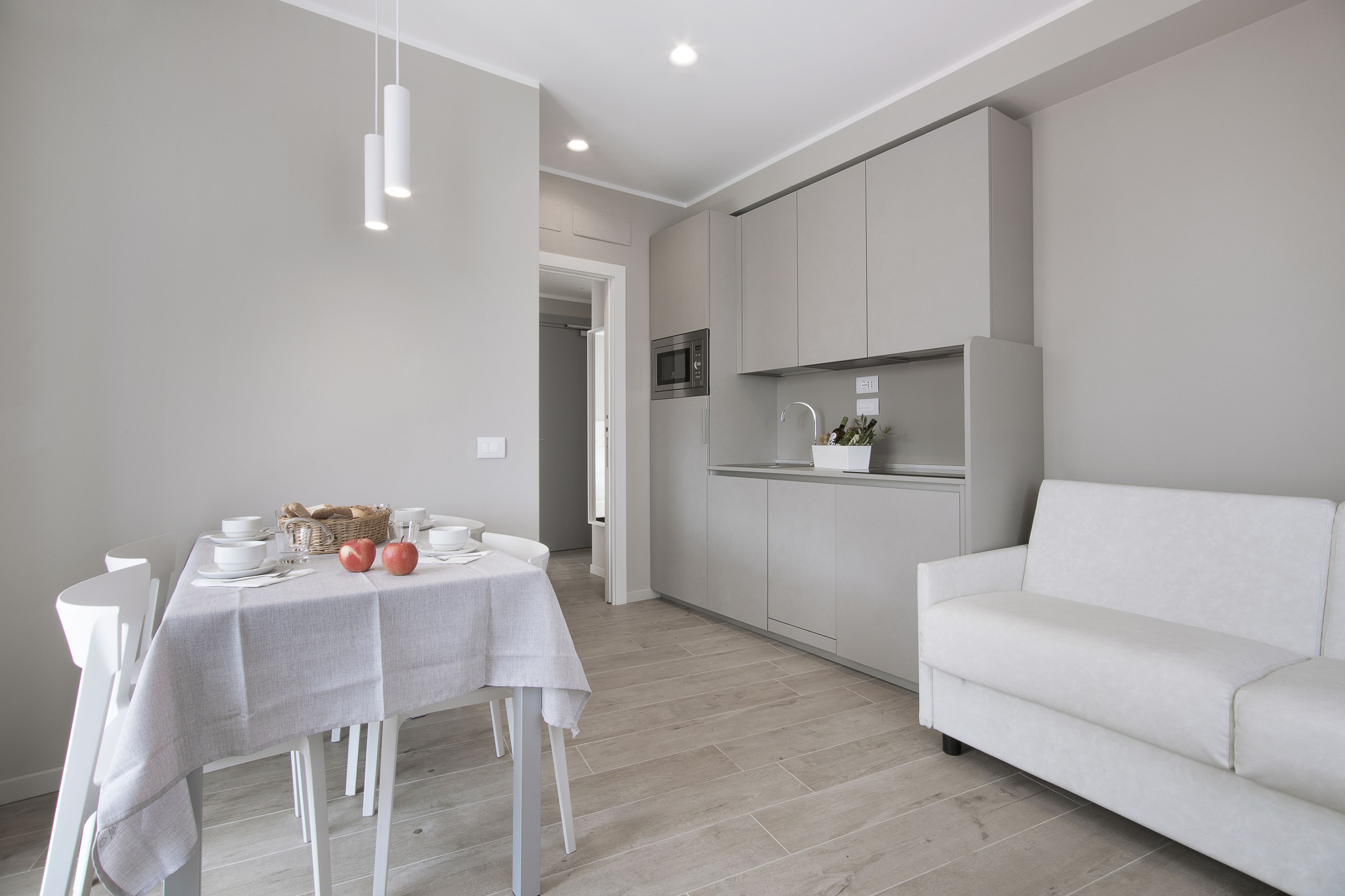 A modern kitchen with white cabinets, a microwave, and a sink. There is a dining area with a table set for two, white chairs, and a white sofa.