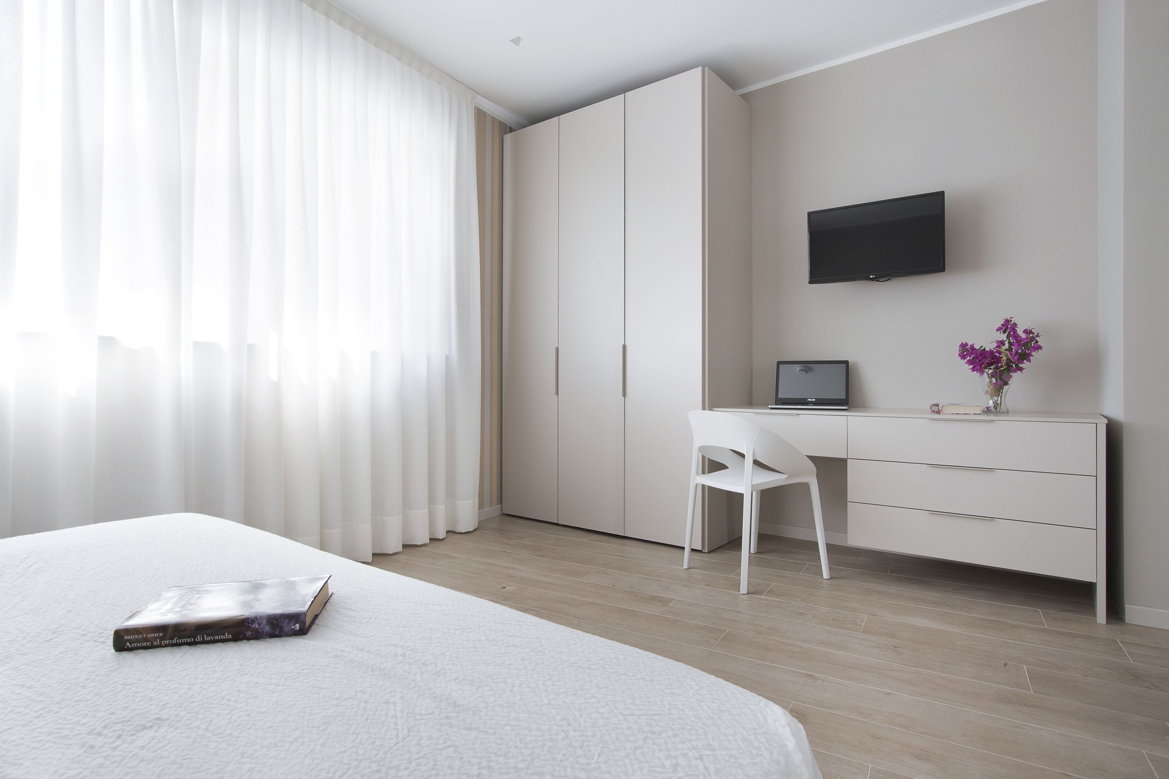 A modern hotel room with a large bed, a desk with a chair, a wardrobe, a TV, and a vase with flowers.